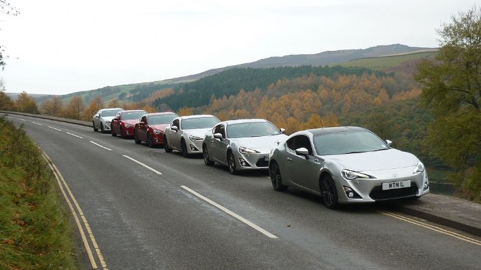 A group of cars parked on the side of the road - Pistonheads - The image shows a long line of sports cars parked on the side of a winding mountain road. The cars are facing the viewer, and each one has a unique body type and color. The vibrant colors of the cars contrast beautifully with the natural surroundings, creating a picturesque scene. The setting gives an impression of a vehicle test route amidst scenic hillside, suggesting that these cars are being exhibited or demonstrated.