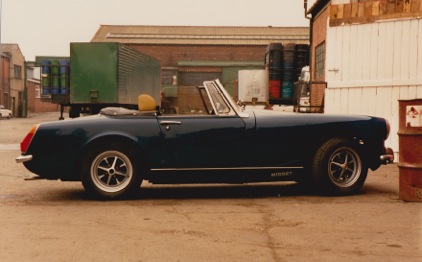 MG Midget Barn Find - Road rally project - Page 1 - Readers' Cars - PistonHeads - The image depicts a classic convertible car parked on a street. The car is a striking blue with a black soft top that is folded down. It features a distinctive white stripe running along the side, and two round headlights are visible. There is a sense of nostalgia in the photo, likely enhanced by the warm tones and sepia effect. In the background, there is a nondescript building and a truck parked nearby. The overall impression is of a casual, non-commercial setting, perhaps in a residential area.