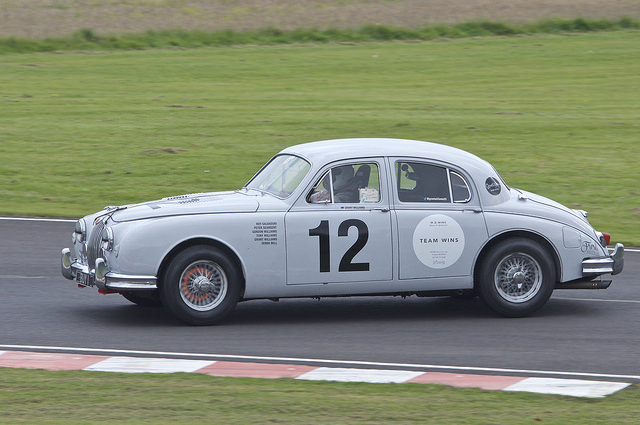 Photography at Castle Coombe - Page 1 - South West - PistonHeads - The image shows a vintage grey race car, likely an Audi Ratzeburg, on a race track. The car is marked with the number "12" prominently displayed on its side and white body, and has "Team Wiz" written in black on the side. The vehicle is powered by a rear-mounted 5.7 liter V-8 engine, which adds to its robust appearance. The car's design and brand suggest it is a racing edition prepared for competitive events. It appears the car is in motion, possibly returning to the pits after a race.