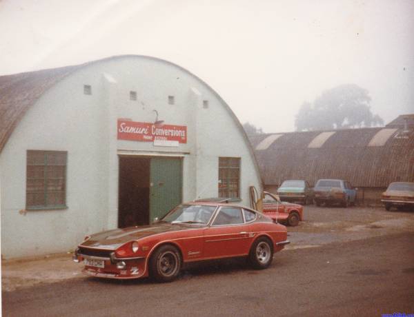 any 240z owners here? - Page 1 - Jap Chat - PistonHeads - The image appears to be a vertical photograph showcasing a vintage-style car, specifically a convertible, parked on what seems to be a lot near a garage with a curved roof. The car has a red leather-topped interior and is designed in the image with a two-door style and a prominent hood scoop. The environment is characterized by a cloudy sky, and there are other vehicles seen in the background. On the side of the garage, there's a red sign with white text that reads "Sam Nawa Conversions." The image has a vintage feel, suggesting it might be an old photo.