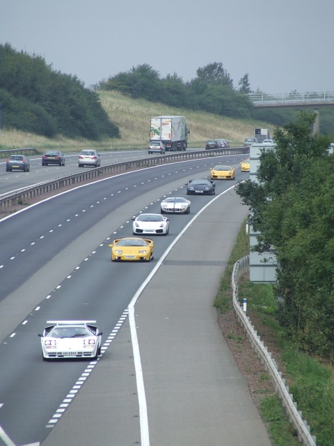 Countach  - Page 6 - Lamborghini Classics - PistonHeads - The image is a color photograph of a wide highway with multiple lanes in each direction. One lane appears to be reserved for construction travel. The roadway itself has a gray surface, with white lines that mark the lanes. In each lane, there is traffic, consisting of various types of vehicles such as cars and buses, indicating a busy time of day. To the right of the highway, there is a grassy median and a guardrail. The sky is overcast, and the lighting suggests either morning or late afternoon. There are no visible incidents or hazards on the road, and the surrounding areas appear to be a mix of grass, trees, and some vegetation.