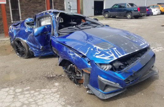 Will the motor/trans have survived this? - Page 1 - General Gassing - PistonHeads - The image shows a heavily damaged blue sports car parked on an unpaved surface. The vehicle is severely wrecked, with its roof and bodywork crumpled, and it has been abandoned in a somewhat disordered manner, likely after an accident. The surrounding area includes a rustic building and a few other vehicles visible in the background. There is no visible text or branding in the image.