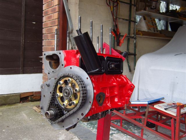 pics - Page 1 - Classic Minis - PistonHeads - The image showcases a close-up view of a transmission of a vehicle. The transmission is red and metallic, with components such as a plastic cover, gear linkages, various bolts, and a metal casing visible. It appears in front of an old car, with tools and a table in the background, suggesting a garage or workshop setting.