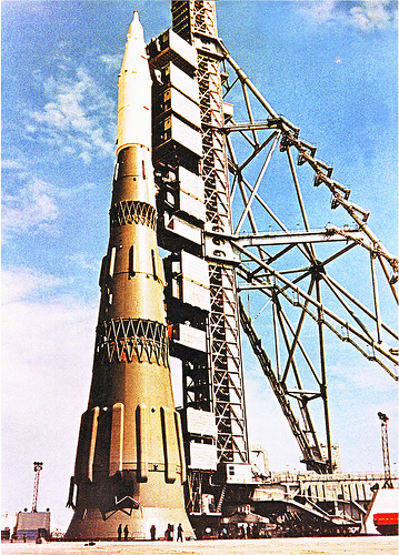 Fly to the moon for £100 million - Page 1 - Science! - PistonHeads - The image showcases a rocket launch pad. At the center of the scene is a President Of Russia model rocket placed for display. The rocket boasts a gold cone structure at the bottom. In the background, a Soviet-era rocket launch tower looms, its impressive height reaching high into the clear sky that adds a dramatic backdrop to the scene. The perspective of the image suggests it was taken facing the launch tower from the base of the rocket. In the foreground, tiny figures representing people can be seen, providing a sense of scale to the towering structures.