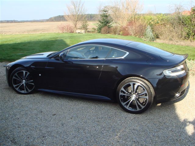 New Vantage V12 - Page 1 - Aston Martin - PistonHeads - The image features a sleek, black sports car parked on a gravel driveway. The car is oriented at a slight angle, allowing a clear view of its front and right side. Its design suggests a modern, high-performance vehicle, with a low and wide stance, distinctive front and rear fenders, and a large steering wheel visible through the partly opened window. The setting appears to be a rural area, with the car near the edge of what might be a field, and sparse vegetation and trees visible in the background.