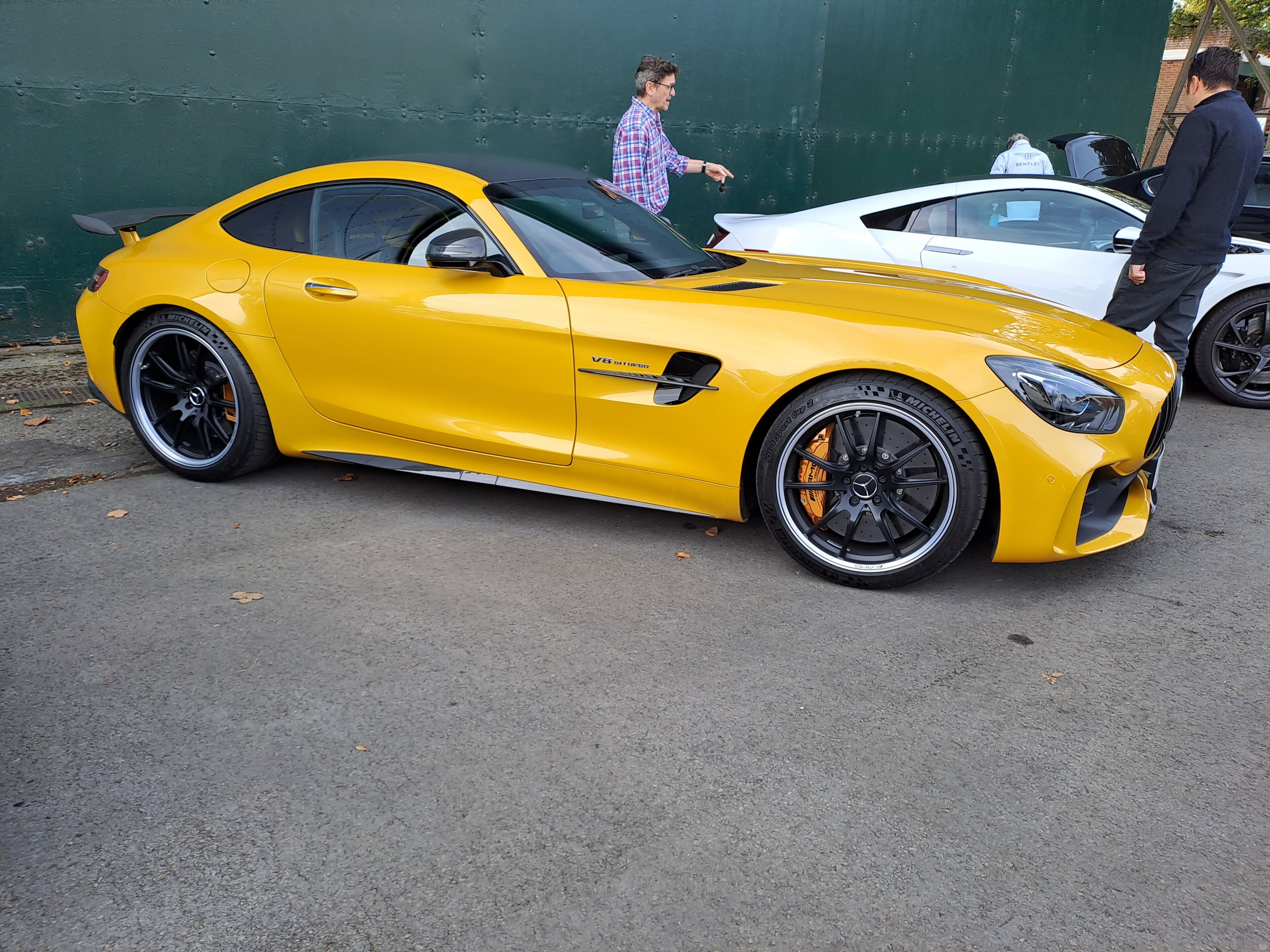 Pistonheads - The image features a yellow Mercedes sports car parked in front of a green wall. The car is sleek and modern, with the Mercedes-Benz logo visible on its side. It has a large rear wing that suggests performance or racing modifications. The tires are low profile, which further indicates the car's sporty nature. In the background, there are two people standing by another sports car, partially obscured behind the first car. The setting appears to be an outdoor parking area during the daytime.