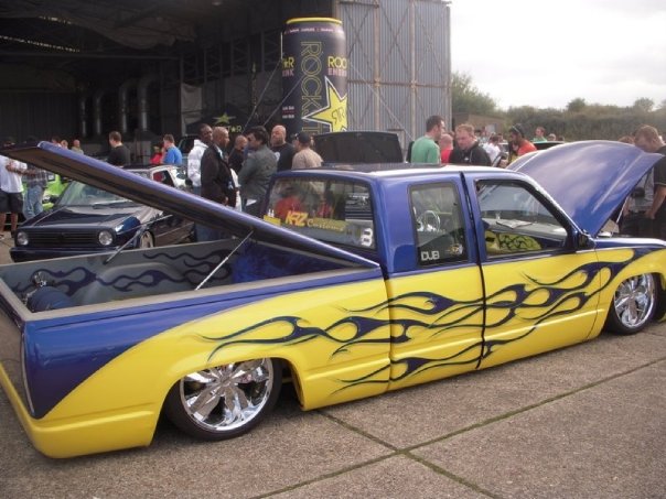 Truck Pistonheads Chevy - The image captures a lively scene at a car show. Dominating the frame is a striking blue and yellow pickup truck, its eye-catching design partially opened to reveal an intricate tattoo-like pattern. The truck's bed, too, is a canvas of bold colors and detailed imagery, adding to its allure.

Around the truck, several people can be seen, their attention drawn to the vehicle. Some are standing closer to the truck, perhaps due to the driver's side door being open, while others are spread out a bit further in the background. Their expressions and body language speak volumes about their interest and admiration for the truck and its distinct modifications.

The image conveys a sense of excitement and appreciation for automotive artistry, as well as the community aspect of car shows. It's a snapshot of a moment that showcases individuality, beauty, and engineering in the world of personal transportation.