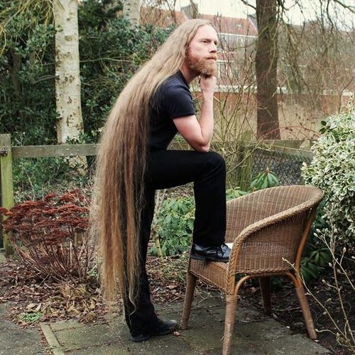 A woman sitting on a bench in a park - In the image, a man with long hair and a beard is leaning against a wooden bench in what appears to be a residential garden. He's wearing a black t-shirt and has his arms crossed. The man's pose suggests he's deep in thought or perhaps contemplating something. Behind him, there's a neatly trimmed lawn with trees visible in the background, suggesting a peaceful suburban setting. The bench is situated on a path, which appears to be a part of the garden landscape.