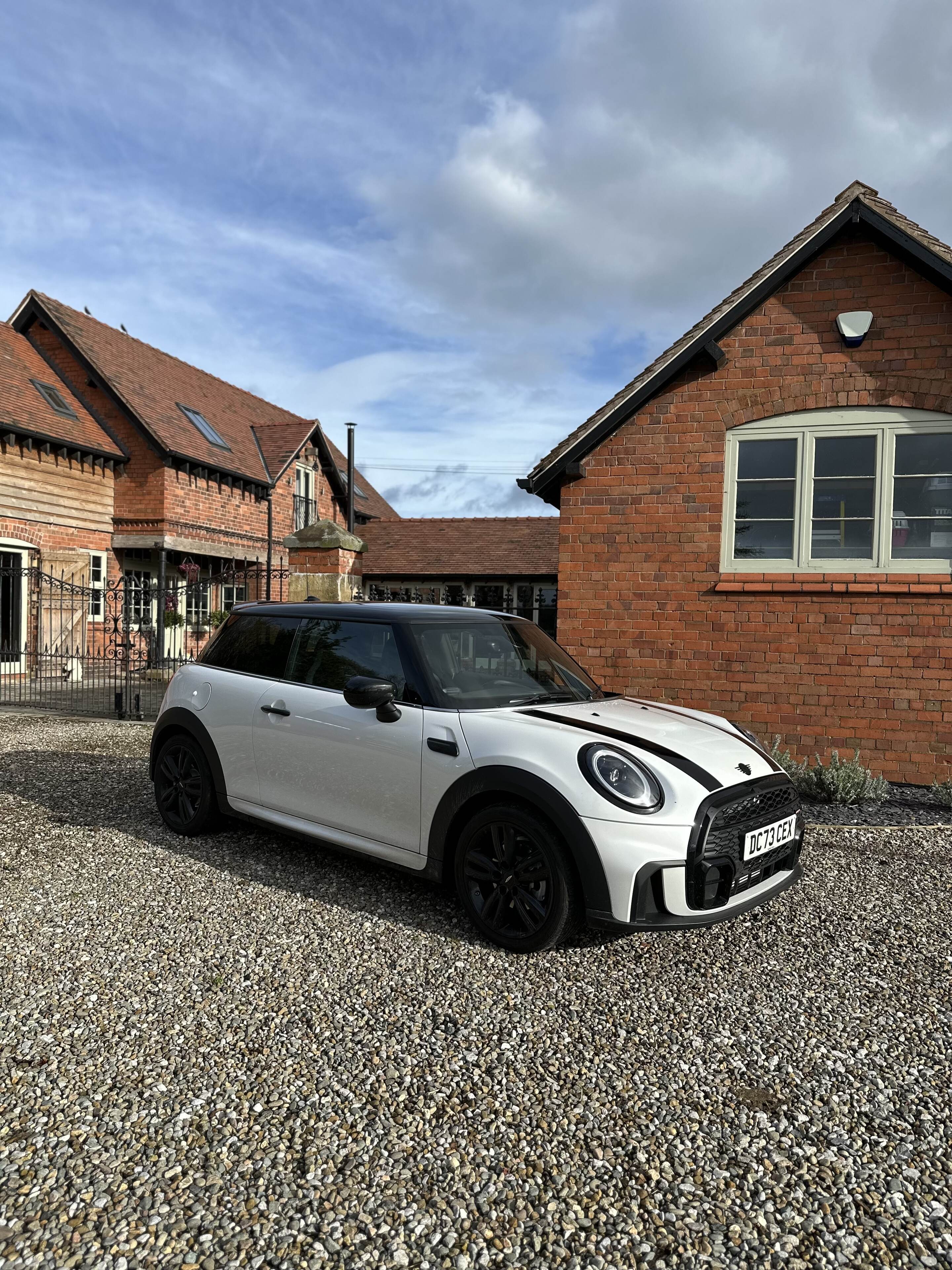 Pistonheads - The image shows a small black sports car parked on a gravel driveway. In the background, there are two-story houses with visible windows and brick chimneys. The sky is overcast with gray clouds, suggesting an overcast day. To the right of the vehicle, there's a garage with a white door. The style of the image is a real estate or lifestyle photograph, capturing the car in its surroundings and providing context about where it might be located.
