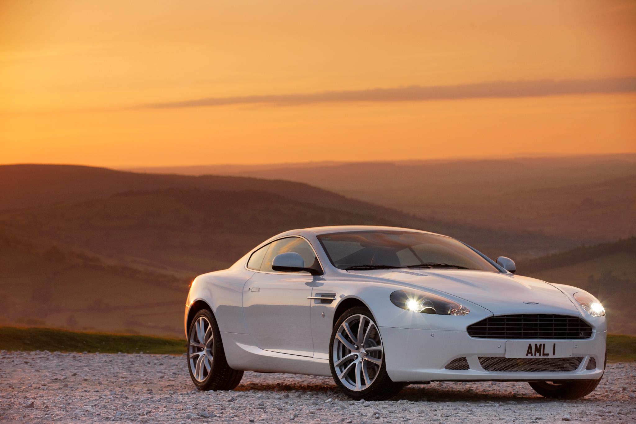 'AML 1' - Page 1 - Aston Martin - PistonHeads - The image showcases a white sports car, prominently displayed in the foreground with a clear view of the license plate reading "AML1". The vehicle is captured at a slight angle, highlighting its sleek design and large alloy wheels. The setting is a scenic sunset view with a beautiful landscape of rolling hills and a dramatic sky filled with orange hues. The car's reflective surface mirrors the sunlight, creating stunning effects with the surrounding environment.