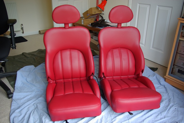 Cerb Seats in an 'S'? - Page 3 - S Series - PistonHeads - The image depicts a room where two red leather car seats have been placed on a protective white tarp. The car seats are positioned facing each other as if they are facing chairs, creating an interesting and unusual setting. On either side of the car seats, there are black office chairs, and in the background, there's a glimpse of a white door and a desk with some papers on it. The room appears to be a personal space or home office, as indicated by the furniture and items around.