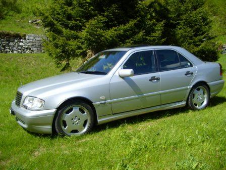 Ticino - Page 1 - Switzerland - PistonHeads - The image shows a metallic gray Mercedes-Benz car parked on a grassy field with green vegetation in the background. The car is four-door, possessing distinguishing features such as a sunroof and a large grille that are typical for Mercedes-Benz models. The vehicle appears to be fairly new, suggesting it might be well-maintained. The background further indicates a rural or semi-rural setting, characterized by trees and a gray gravel road.