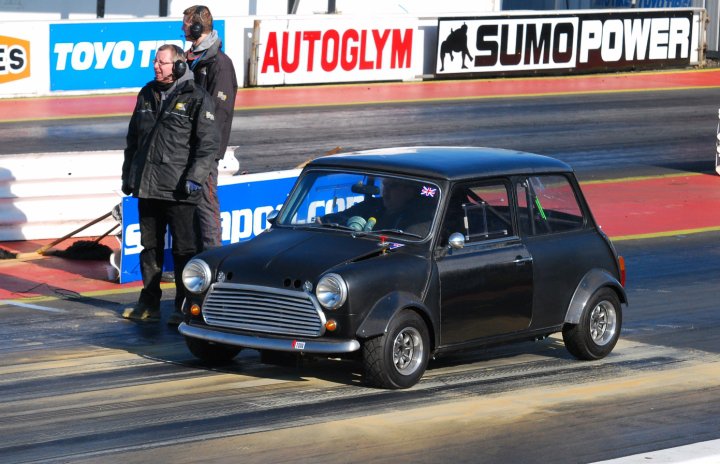 Anyone know this car? - Page 1 - Classic Minis - PistonHeads - The image shows a small, black Mini Cooper car on a race track. There's a man standing in front of the car, wearing a black coat and a black helmet, possibly preparing to drive the car. In the background, there are banners with various logos, including one that reads "Stunt Power". The overall scene suggests that this might be a motorsports event that involves stunts or demonstrations.