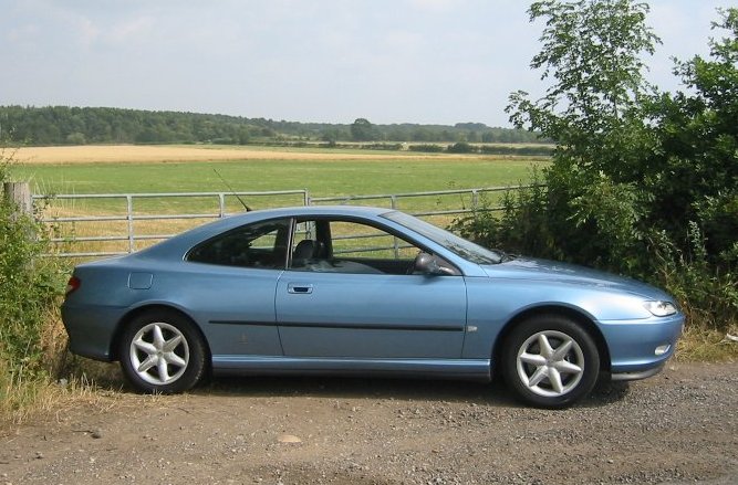 Pistonheads Altering Wheel Pcd Alloy - The image features a blue compact sedan parked on a dirt road adjacent to a wire fence. The car is positioned with its side profile facing the camera, emphasizing its design and a glimpse of the surrounding country landscape. The terrain is flat and open with a distant view of the countryside. Vegetation, including bushes, lines the fence, adding a touch of nature to the scene. The afternoon sun casts soft lighting on the car, enhancing its color and reflecting off the ground near the tire.