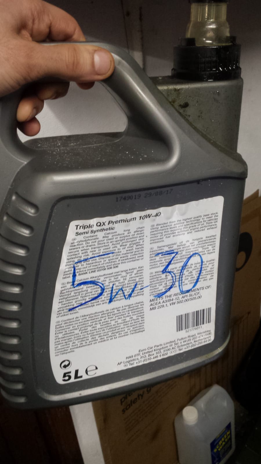 Pistonheads - The image shows a person's hand holding a plastic container labeled "5W-30". The label includes a barcode and some text, but the details are not fully visible. In front of the container, there is a white piece of paper with black lettering indicating that the oil should not be mixed with gasoline. The setting appears to be an indoor environment, possibly a garage or workshop, suggested by the presence of a cardboard box and what looks like a cleaning product in the background.