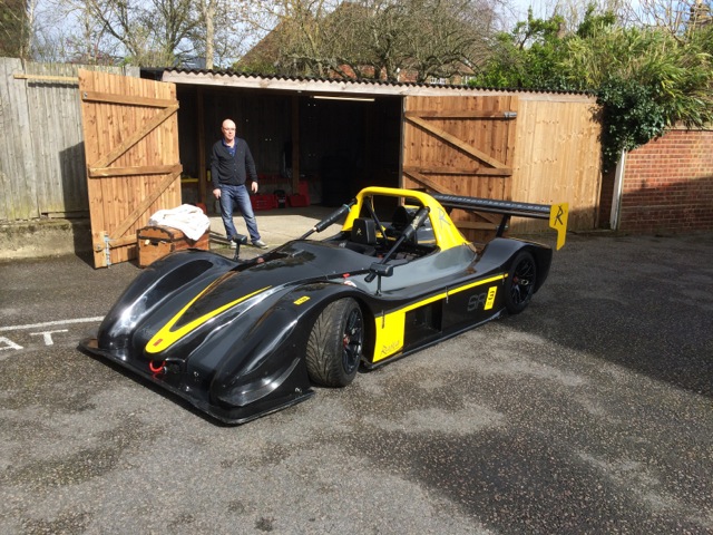 A motorcycle is parked in a parking lot - Pistonheads - The image shows a man standing next to an unusual sports car. The car is striking due to its black and yellow color scheme and its streamlined design suggests high speed. It appears to be parked in an outdoor setting, possibly in front of a garage. The man, who is not the main focus of the photo, is standing slightly behind and to the left of the car. The environment suggests a garage setting with a paved parking lot.