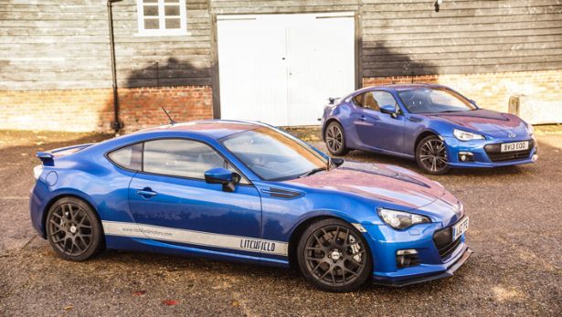 A blue car parked in front of a building - Pistonheads - The image features two identical cars parked side by side on a gravel surface. These cars appear to be sports cars judging by their sleek design and distinctive front indicators. They are of a similar make and model, suggesting they may belong to the same model line. The color of both cars is a striking two-tone blue with the top half being lighter blue and the bottom half being darker blue. Their placement outside a building imparts a sense of motion and dynamic style to the scene.