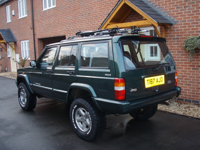 Jeep Cherokee Pistonheads Lift - This image features a green SUV parked on a paved driveway next to a brick house. The vehicle has a spare tire mounted on top of its roof rack, and a ski rack is visible above the car. A red reflector is affixed to the back of the car, providing visibility, and there's a yellow license plate on the back bumper. The overall scene suggests a residential neighborhood, and the weather appears to be clear and sunny.