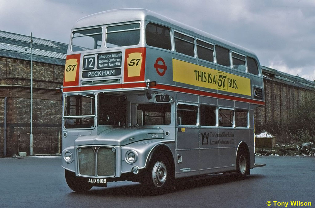 A red double decker bus driving down a street - Pistonheads - This image features a classic double-decker bus, predominantly painted in silver, that stands out against a backdrop of a run-down building and another building under construction. The bus bears the numbers "57", along with the sign "Peckham" on its front. The bus has an additional sign on the side that reads "This is a 67 bus." It's driving on a road next to a chain-link fence. This image portrays a scene of urban transportation and urban decay coexisting.