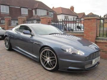 Pistonheads - The image features a two-door sports car parked on a brick driveway in front of a residential house with red brick exterior walls and white lettering on the side. The vehicle is a dark gray or possibly black color, possibly a luxury or exotic sports car given the design cues. There are no visible texts on the car itself. The sky in the background is overcast, suggesting that the photo was taken on a cloudy day.