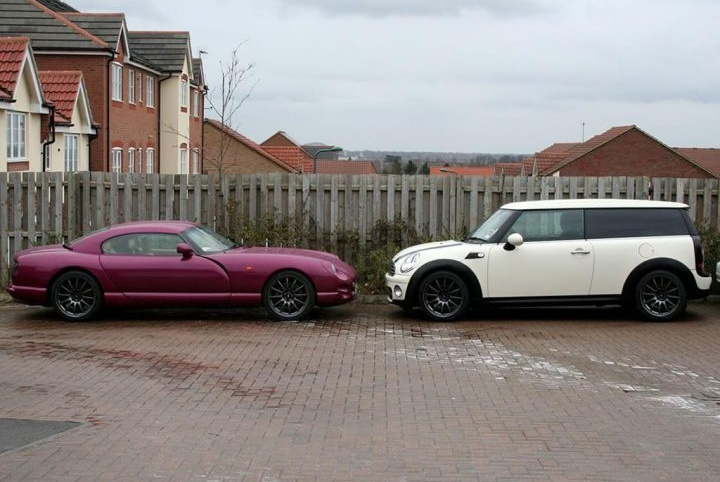 A car parked in a parking lot next to a car - Pistonheads - The image shows two vehicles parked next to each other on a wet paved area. To the left is a maroon luxury sports car, known as a Maserati Ghibli, characterized by its sleek design and large alloy wheels. To the right is a white Mini Cooper Clubman, smaller in size and also having a distinctive design with a door at the rear for additional cargo space. Both cars have alloy wheels, and the wet ground reflects the sky above. In the background, there are residential buildings, suggesting this parking area is in a residential or suburban neighborhood.