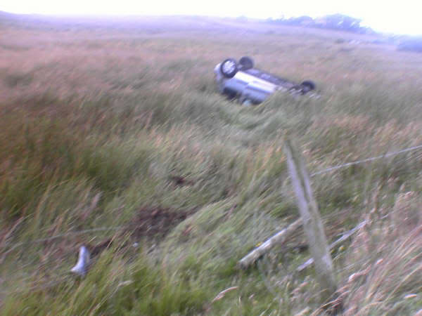 Most bizarre and/or unlucky accident outcome encountered. - Page 1 - General Gassing - PistonHeads - The image shows a scene of a car flipped over onto its side in a grassy field. The car is silver in color and its rear is both crushed and scraped. Its wheels are missing, adding to the sense of a significant accident or impact. There is green foliage surrounding the vehicle, and the car's position suggests it has rolled extensively or been impacted forcefully. The sky appears to be overcast, providing a somber backdrop to the scene.