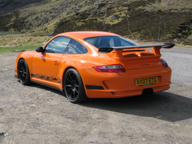 I'm not a Porsche man but.. - Page 1 - Noble - PistonHeads - The image shows an orange race car parked on a dirt surface. The car appears to be a sports or race car, likely a Porsche given its design cues. The rear wing of the car is prominent, suggesting it is designed for aerodynamic efficiency. The car is bathed in sunlight, casting a warm glow over its surface and enhancing the orange hue. The background features a rocky hillside with sparse vegetation, adding a natural element to the composition.