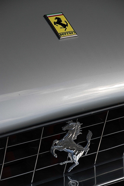 Goodwood Breakfast Club, Pre 1966 classic sunday - Page 1 - Goodwood Events - PistonHeads - The image shows the hood of a car with a Ferrari logo centrally displayed. The Ferrari logo is a stylized horse with a raised leg, indicative of the Ferrari brand. The background of the image is a plain, untextured, metallic gray. The reflection on the hood suggests that the vehicle is metallic, reflecting the vehicle's colors. This is a photograph of a Ferrari vehicle, and the angle gives a view that also reflects the interior of the vehicle on the open hood.