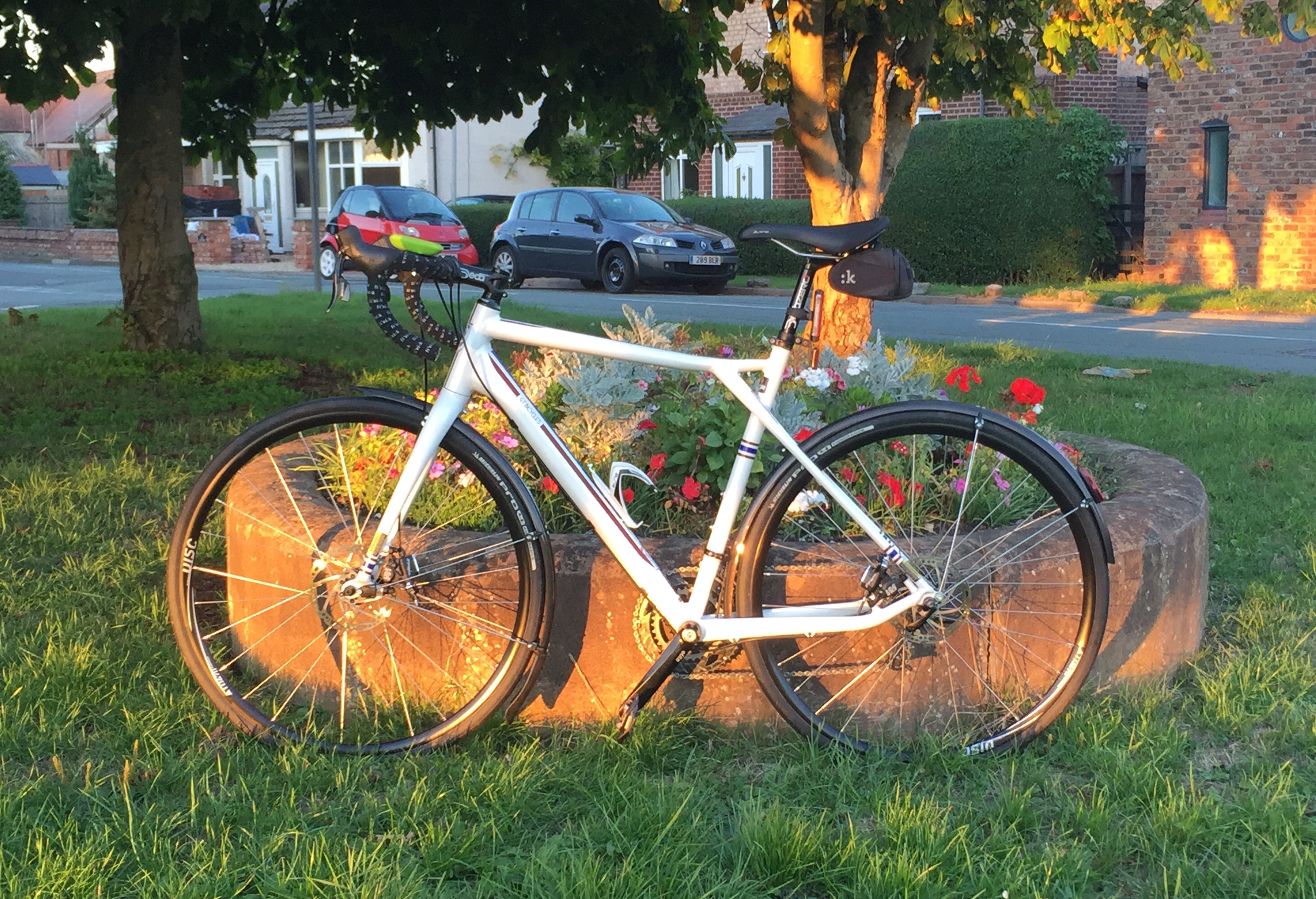 The "Photos From Today's Ride" thread... - Page 398 - Pedal Powered - PistonHeads - The image shows a white bicycle parked leaning on a stone retaining wall. The bike is equipped with black tires and rims, and it is well-maintained, with visible gears and a thin lexicon cable running up the back of the chainstays. In the background, there are parked cars, including a red hatchback and a black sedan, suggesting a residential area. The setting appears to be a well-kept garden or park, with green grass and various plants and flowers enhancing the overall bucolic atmosphere.