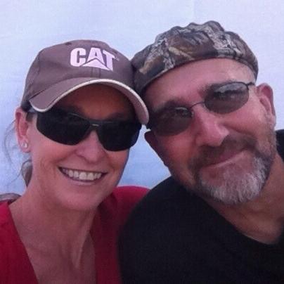 A man and woman pose for a picture - The image is a photograph of two adults, a young woman and an older man, standing next to each other with their heads together. Both individuals are wearing sunglasses and matching style accessories, with the woman wearing a cap and the man a baseball-style hat. They appear to be in a positive mood, as they both grin towards the camera. The man has a casual, yet well-groomed look with facial hair, while the woman has short hair framed by glasses. They are outdoors, as light reflects off their sunglasses and the background is not distinct. The overall impression is of a joyful moment captured between the two individuals.