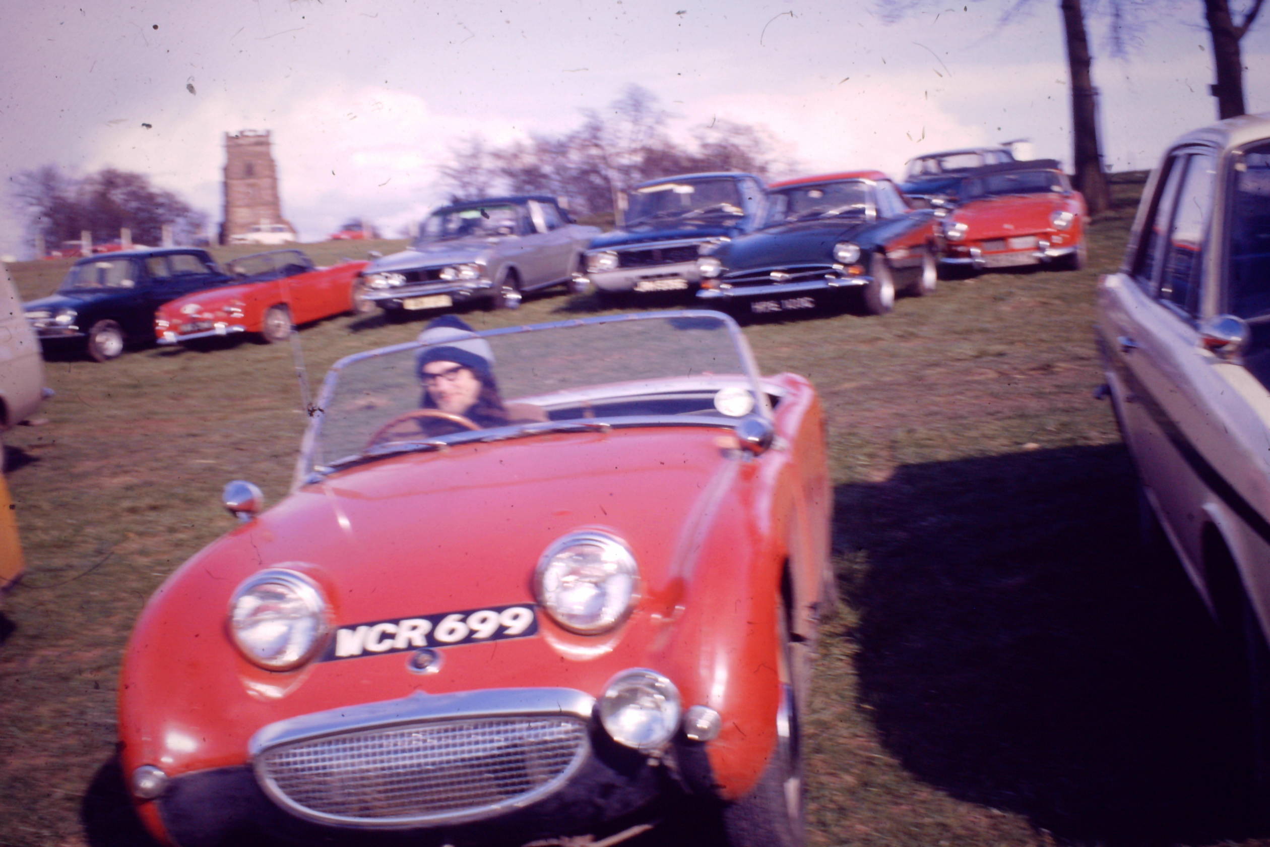 Longest ownership and still yours - Page 3 - Classic Cars and Yesterday's Heroes - PistonHeads - In the center of the image, a vintage red sports car is parked on grass, surrounded by other classic cars. A person stands outside the red car, their attention seemingly captured by something off-camera. The setting appears to be an outdoor event or gathering related to classic cars. The cars are arranged in a line, with the red sports car being the centerpiece. The vehicles range from older models to more modern classics, creating a vibrant display of automotive history.