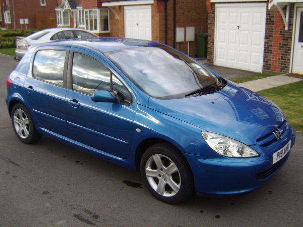 My Car - Page 1 - Readers' Cars - PistonHeads - The image shows a sleek, dark blue Honda car parked on the side of a road. The car appears to be a 4-door model and is positioned facing the camera, giving a clear view of its front. In the background, there is a residential street with a house on the right side of the image and another vehicle visible in the distance. The sky is partly cloudy, suggesting it might be a mild day. The overall scene conveys a sense of ordinary urban life.