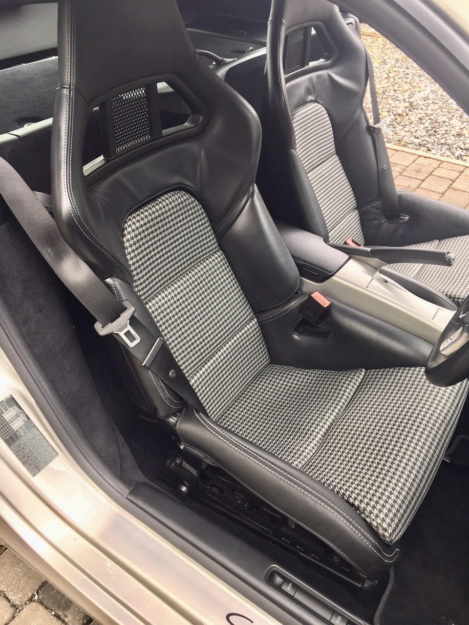 Pistonheads - The image showcases the interior of a sports car, specifically focusing on its driver's seat. The seat is adorned with a black and white checkered pattern, indicative of a racing or rally theme. The material appears to be suede, adding to the sporty aesthetic of the vehicle. 

The car is currently parked indoors, as evidenced by the white wall in the background and a section of wooden flooring visible on the left side. A black seat belt is prominently displayed across the middle of the seat. The overall setting suggests that the car might be a showcase or a model for potential buyers, given its pristine condition and meticulous arrangement.
