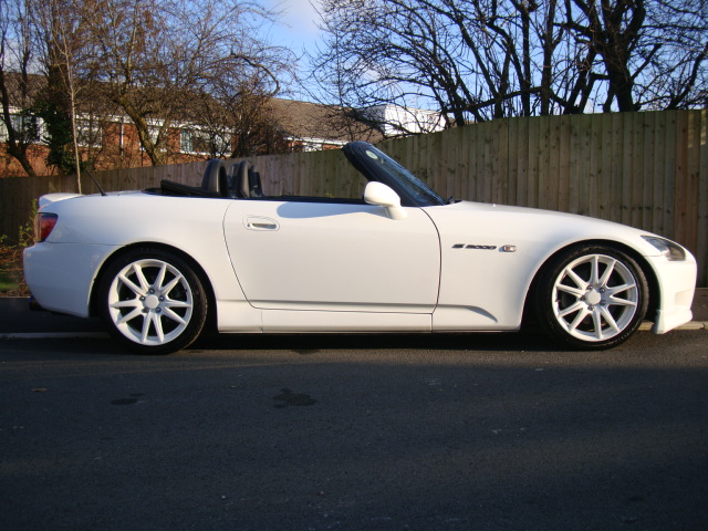 Pistonheads - The image features a white sports car parked on the side of a road. The car is sleek, with visible aerodynamic enhancements like a large spoiler. The wheels are white with a silver rim, adding to the car's sporty appearance. The car is positioned on a paved surface, adjacent to a wooden fence, and a few trees can be seen in the background, suggesting a suburban setting. The sky is clear, indicating good weather conditions.