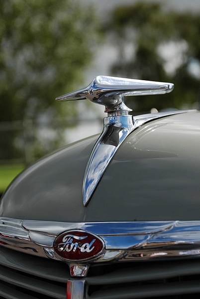 Goodwood Breakfast Club, Pre 1966 classic sunday - Page 1 - Goodwood Events - PistonHeads - The image features a close-up of a vintage car's hood ornament. The ornament is a polished, silver-colored piece shaped like a V, with a curved base that mounts on the Ford trademark emblem located in the center of the grill. The surface of the ornament reflects the overcast sky and some tree branches behind it, giving it a distinct shine. The car itself is gray but its surface tone reflects the changing sky conditions. There is no visible text on the image to provide additional information. The style of the photograph captures the details and texture of the metal ornament clearly.