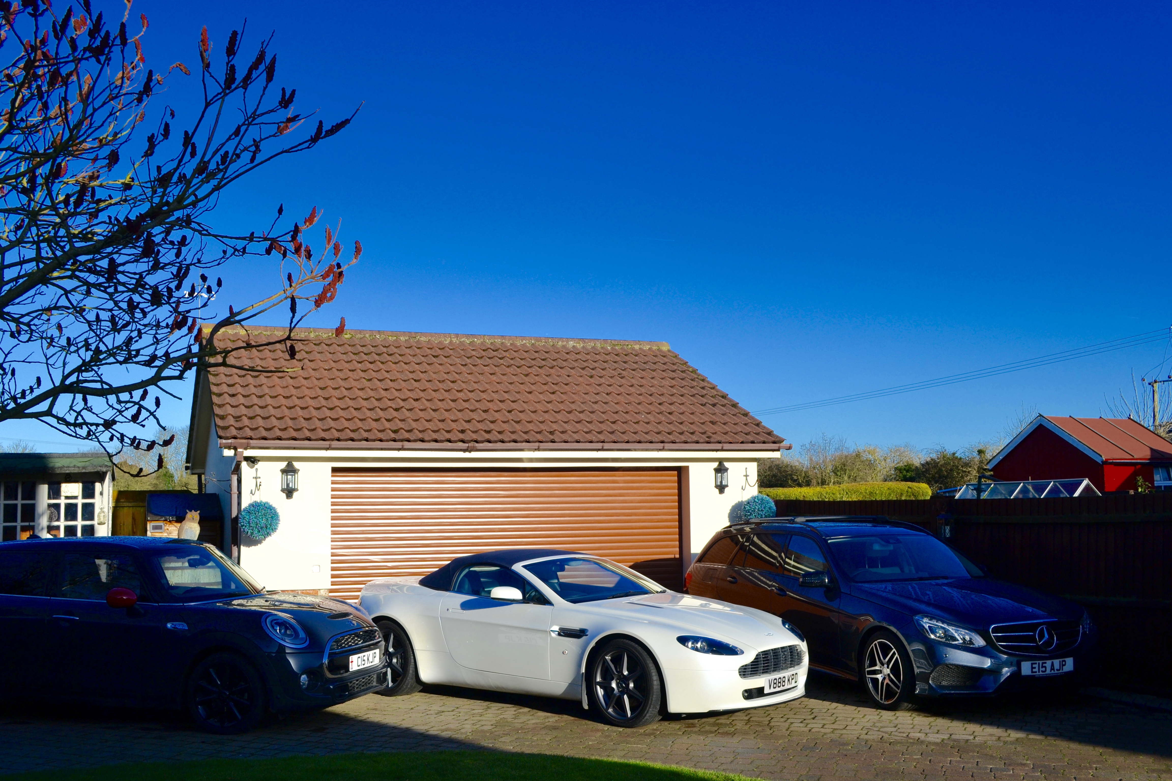 How about an Aston photo thread! - Page 138 - Aston Martin - PistonHeads - The image presents a serene scene from a residential area. Two cars are parked side by side in front of a garage, one of which is a striking white, and the other is a sleek black. The garage they're parked in has a red roof, adding a pop of color to the scene. The sky overhead is a clear blue, suggesting that it might be a sunny day. The overall mood of the image is calm and domestic.