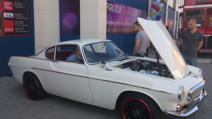 Camberley Town Centre car show - Page 1 - Thames Valley & Surrey - PistonHeads - The image features a vintage white car prominently displayed with its hood open, revealing a shiny silver engine. A group of onlookers is interested, and two gentlemen are standing close by, examining the car and its engine. The car appears to be of classic design, and its surroundings hint at an urban setting with blue walls in the background. The overall atmosphere is one of curiosity and appreciation for the vehicle.