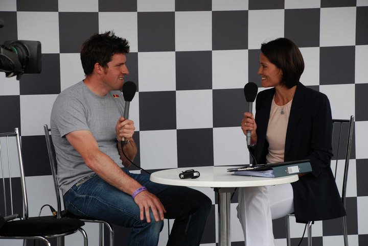 Post your Pics, Celebrities at  FOS 2011 - Page 1 - Goodwood Events - PistonHeads - In the image, a man and a woman are seated at a checkered table in what appears to be a broadcast or interview setting. The man is holding a black microphone, suggesting he is the one conducting the interview. He is also wearing a bracelet. The woman is smiling and seems to be on a podcast or a radio show. The table has a few items on it, including a pen and a green folder, indicating some sort of professional discussion. The background features a wall with a grey and white checkered pattern, adding to the professional atmosphere.