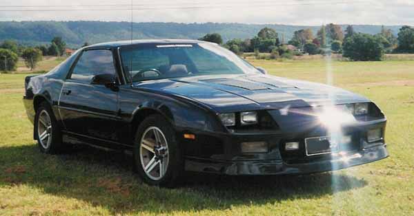 Chevrolet Big Camaro Pistonheads Tavs Iroc - The image features a sleek, black sports car parked in a grassy area. The car is positioned at an angle to the camera, with its front facing the viewer. The car is equipped with a spoiler above the rear windshield and there are black panels over the rear lights. The engine hood has air vents and there is one visible wheel at the front of the car. The setting sun casts a warm, golden glow on the car, emphasizing its shiny surfaces and reflecting patterns of light and shadow. The background shows a spacious, open field under a gradually lightening sky, suggesting either dawn or dusk.