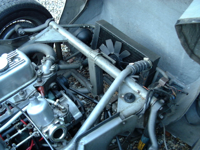 Early TVR Pictures - Page 34 - Classics - PistonHeads - The image shows a close-up view of the underside of a vehicle, displaying parts of the motor and related components. The engine component is dirty and appears to be leaning to the left when viewed from the front. The supporting frame or bracket to which the fan or an electric motor is attached has a metallic, possibly chrome finish, and is connected to what seems to be a large fan or electric component. There are other mechanical parts scattered around, suggesting that the vehicle is currently being worked on or has been recently serviced. The background is blurred, emphasizing the focus on the vehicle's inner workings.