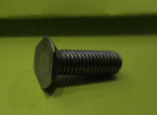 Mystery anti-tamper screw - Page 1 - Home Mechanics - PistonHeads - A single tapered metal screw is visible. The screw appears to be embedded into a green surface. Its head is visible on the left side of the screw. The screw shines, indicating a reflective quality to its metal. The surface beneath the screw is smooth and flat.