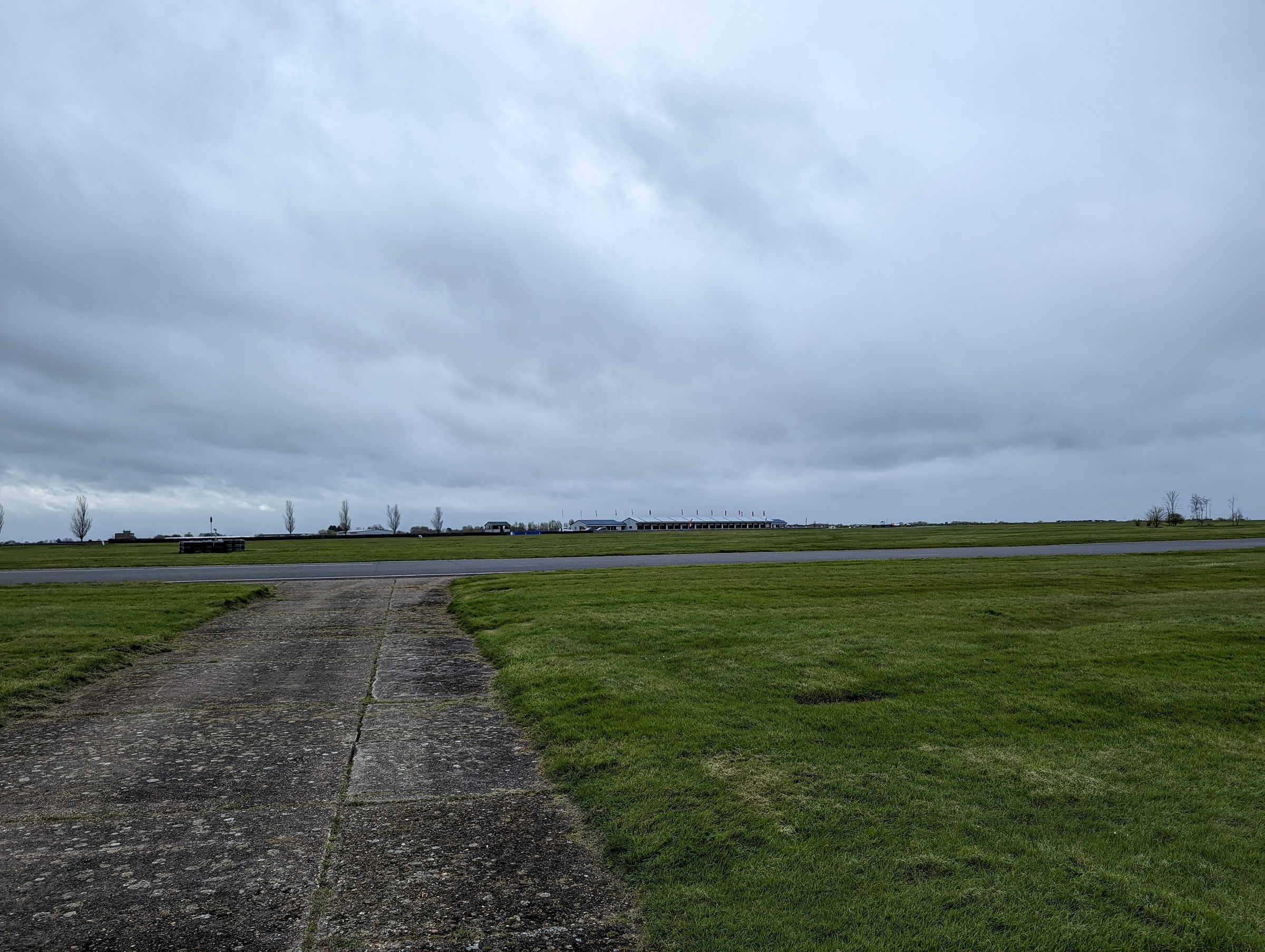 Pistonheads - This is a color photograph showing an overcast sky above a runway with a paved surface. The runway leads to the horizon, where several buildings and structures can be seen under the clouds. In the foreground, there is a grassy area next to a patch of bare earth, indicating a field or a park. There's a watermark on the image that reads "grey" which could suggest the name of the photographer or the software used.