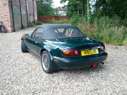 1991 mx5 limited edition- Rare i think pls help? - Page 1 - Mazda MX5/Eunos/Miata - PistonHeads - This image features a Japanese sports car parked outdoors on a gravel driveway. The car has a distinctive black and yellow license plate. It is a two-door, convertible sports car with a visible rear windshield. The vehicle is situated next to a landscaped area with trees and shrubs, and in the background, there is a building with arched doorways.