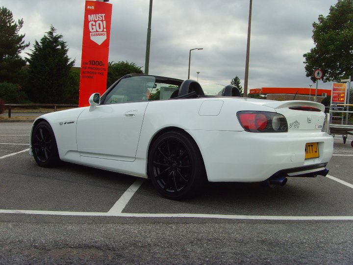 Pistonheads - The image features a parked white sports car in a parking lot. It appears to be a two-door vehicle, possibly a sports car given its sleek design and rims. The vehicle is predominantly white, which is evident against the more muted surroundings. The background indicates that the photo was taken during the day under an overcast sky, and it includes a sign for an electronic product sale. The car is parked near a barrier, and the overall setting indicates an urban environment.