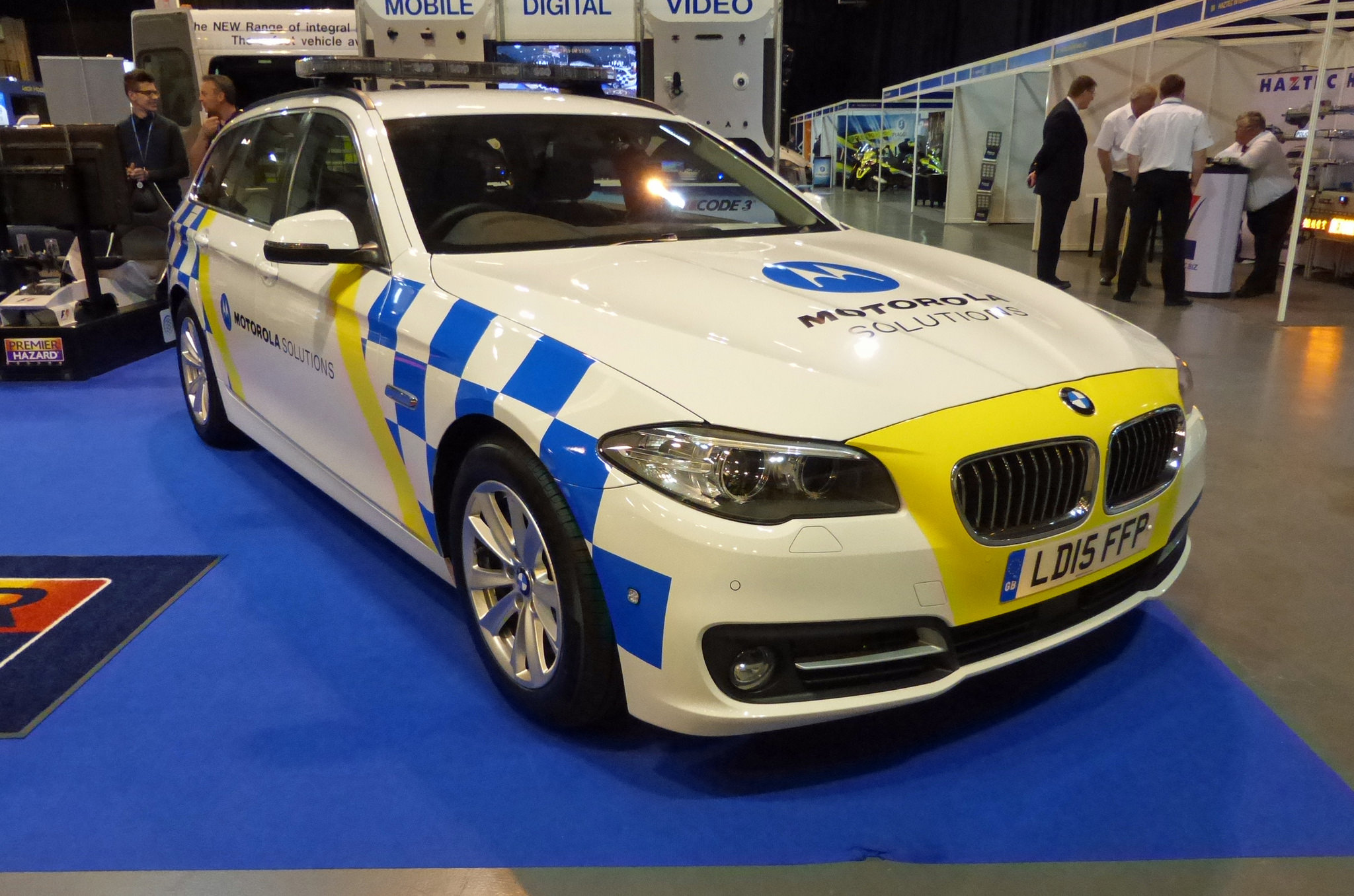 Motorola car trying to imitate police - Page 1 - General Gassing - PistonHeads - The image features a striking hybrid sports car on display. The car's distinctive blue and yellow checkered pattern covers the hood and sides, adding a chunky, racing feel. The brand name "MIRAGLOT" is prominently displayed in white on the side of the car. The license plate reads "LD15 FP," and the car is parked on a blue carpet with a black and gold diamond pattern. The setting appears to be an auto show or similar event, as suggested by the background, where a crowd of people dressed in business attire can be seen walking, standing, and conversing.