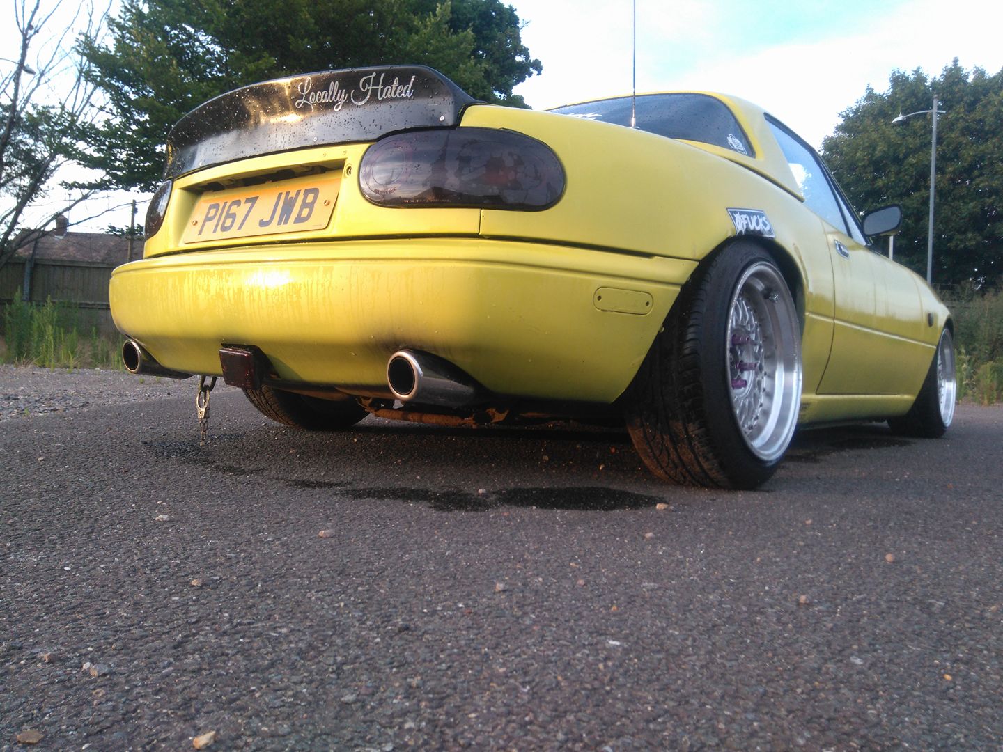 Saved from the "Scene" Mk1 MX5 rescue-ish - Page 1 - Mazda MX5/Eunos/Miata - PistonHeads - In the image, there is a striking yellow car positioned at an angle, showcasing the sun reflecting off its hood. The car has a license plate reading "PIG7JB", and its rear wing bears a contrary message reading "Lovely Hate." The vehicle features a black aftermarket wing and a black and white aftermarket bodykit, adding to its distinctive appearance.