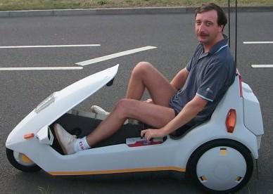 Renault Twizy - Page 1 - French Bred - PistonHeads - In the image, a man is seated comfortably in a small, white, three-wheeled vehicle. He is wearing a blue shirt and his feet are up, relaxed in the pedal car. The vehicle's design is somewhat unusual, with a large opening at the front, making it appear more like a auto -mobile, as shown by the man's pose. The road behind him suggests a journey or adventure ahead.