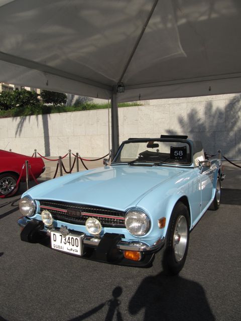 Classic Emaar Pistonheads - The image showcases a classic blue car, likely a Triumph Spitfire, parked under a white tent or awning. The car is adorned with a "No Further" sticker prominently displayed on the windshield. The car has a shiny metallic hood and unique red stripes running along its side. Behind the car, red barriers can be seen, suggesting a level of protection or exclusivity. To the left of the car, another red car is partially visible, partially obscured by the tent's shadow.