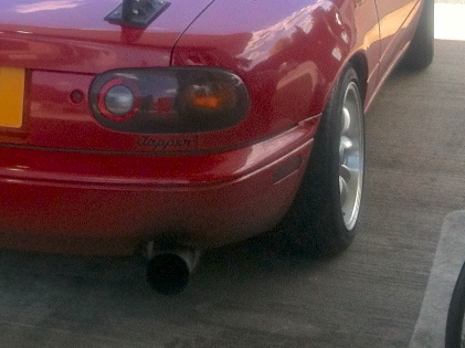 A disturbing sight - Page 1 - Mazda MX5/Eunos/Miata - PistonHeads - The image shows a close-up view of the front end of a red car, featuring its left headlight and part of the tire on the left front wheel. The car appears to be standing still, as suggested by the shadow it casts directly below it. The license plate is mounted on the front bumper, and there seems to be a metal accessory mounted in the space where the side mirror would be on the left side. The background of the image is an indistinct gray surface, possibly a pavement or concrete floor.