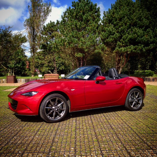 A red and black car parked in a parking lot - Pistonheads - The image shows a vibrant red sports car parked on a black-topped surface characteristic of cobblestone patterns. The car is a convertible style, with the top down, revealing a two-seat interior. It has black rims that contrast with the car's body, and there's a logo on the front that suggests the manufacturer. The setting appears to be outdoors, with lush green trees forming a picturesque background. The vehicle is positioned at a slight angle, allowing a view of one side and the rear end.